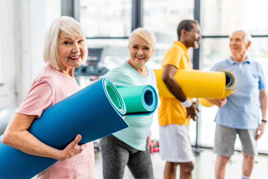 selective focus of senior sportswoman holding fitness mat and her friends standing behind at gym PLACEHOLDER IMAGE